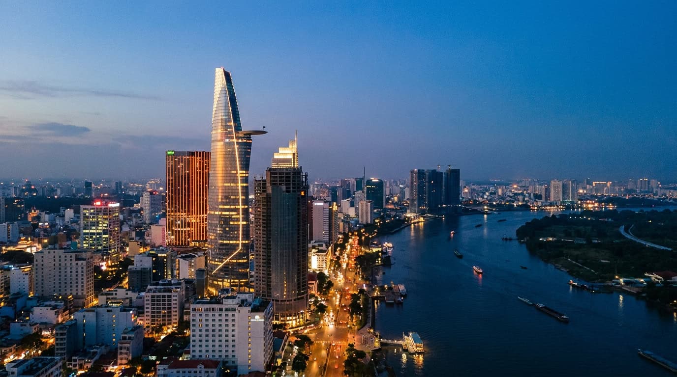 Photography from a Saigon rooftop at blue hour showing the city skyline with Bitexco Tower illuminated, the Saigon River visible in the distance, and a twilight atmosphere over the city.