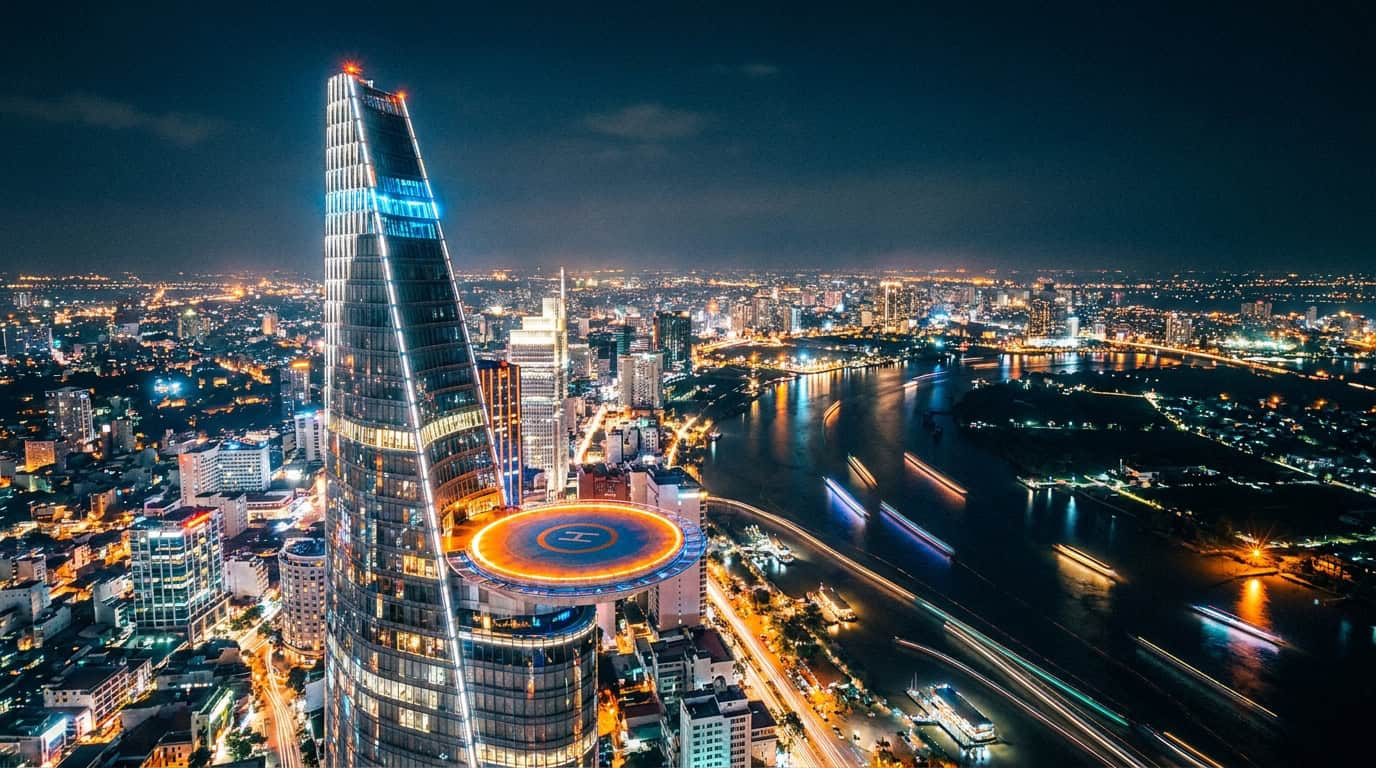 Bitexco Financial Tower illuminated at night with the glowing city of Ho Chi Minh City spread below, District 1 street lights and the Saigon River visible in the background.